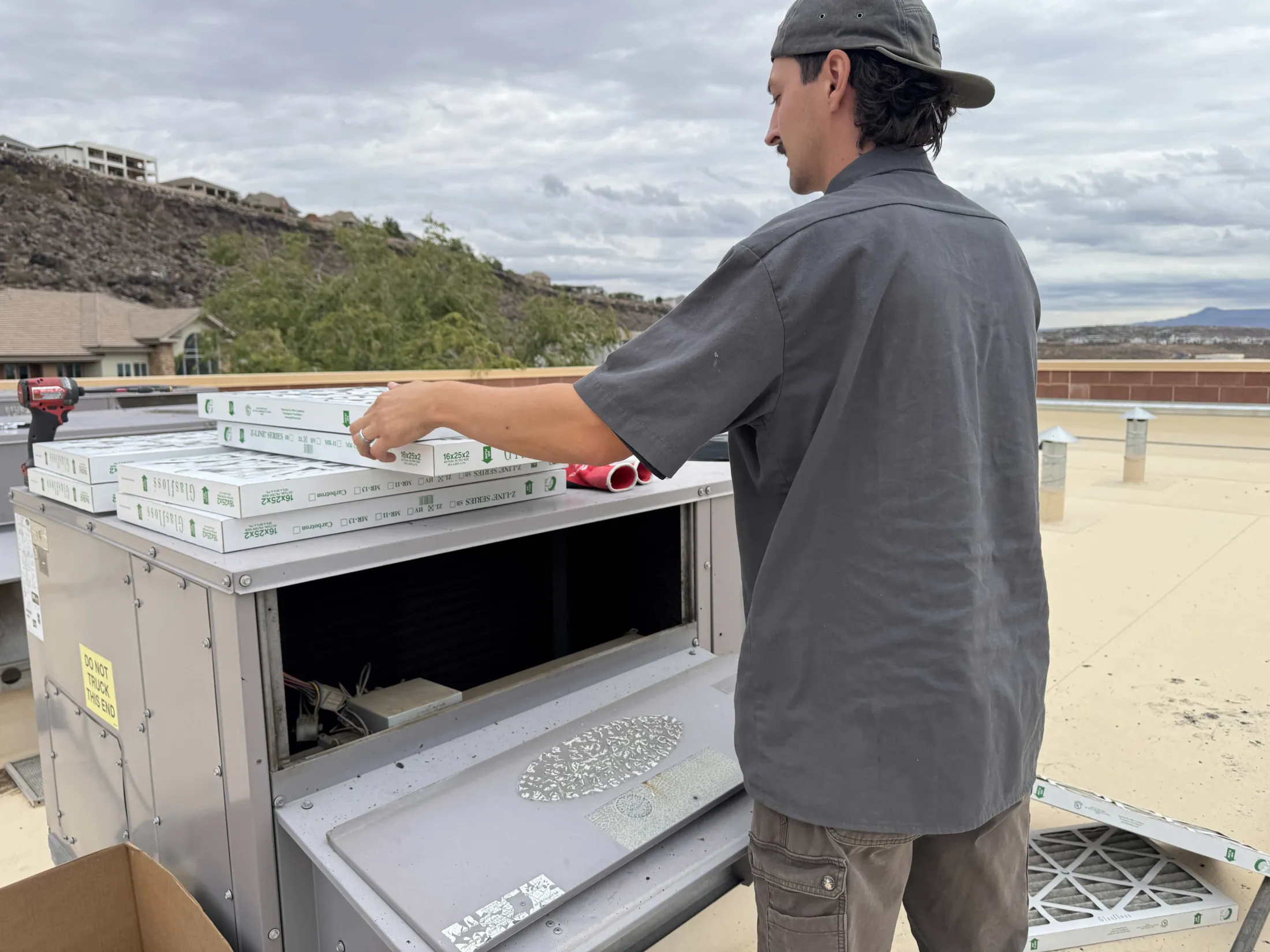 A technician on a rooftop reaches for a stack of new air filters staged on a package unit, with the Red Hills and a residential neighborhood in the background.